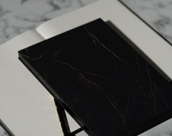 Puede incluir: Un diario negro con diseño de mármol y vetas doradas descansa sobre un cuaderno blanco punteado. La cubierta del diario es negra brillante y las páginas son visibles. El fondo es una superficie de mármol.