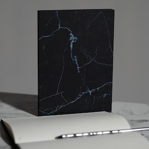 May include: A black marble-patterned notebook stands upright, showcasing a detailed veining design in shades of blue and white. An open notebook with blank pages and a silver pen are in the foreground, suggesting a writing or sketching theme.