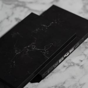 Könnte beinhalten: Ein Stapel aus drei schwarzen, rechteckigen Objekten mit marmorähnlichem Muster. Die Objekte sind leicht angewinkelt angeordnet, wodurch ihre Kanten und die marmorierten Oberfläche darunter sichtbar werden. Die Objekte haben eine matte Oberfläche.