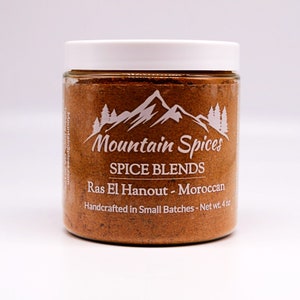 May include: A glass jar with a white lid filled with brown Ras El Hanout spice blend. The label reads "Mountain Spices Spice Blends Ras El Hanout - Moroccan Handcrafted in Small Batches - Net wt. 4 oz".