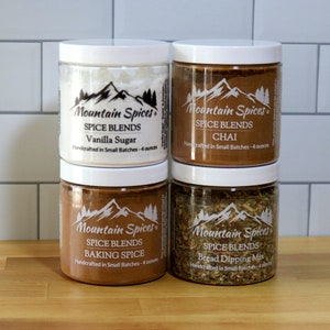 Può includere: Quattro barattoli di vetro con coperchi bianchi contenenti diverse miscele di spezie. I barattoli sono etichettati "Mountain Spices" e elencano il nome della miscela di spezie, "Vanilla Sugar", "Chai", "Baking Spice" e "Bread Dipping Mix". Ogni barattolo contiene 4 once di miscela di spezie.