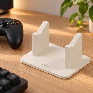 Op de afbeelding: Een witte rechthoekige controllerstandaard met twee verticale steunen met schuine inkepingen voor het vasthouden van een gamecontroller. De basis van de standaard heeft de naam Alex in cursief schrift op de voorzijde van het witte materiaal.