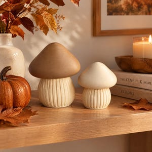 Peut inclure: Deux figurines décoratives en forme de champignon avec des chapeaux lisses et arrondis et des pieds à côtes verticales. Le plus grand champignon a un chapeau beige, tandis que le plus petit a un chapeau crème. Les deux pieds sont de couleur crème.
