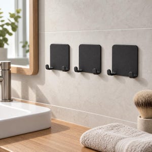Puede incluir: Tres ganchos de pared cuadrados en negro mate, cada uno con dos puntas curvadas hacia arriba. Estos ganchos de tipo adhesivo están montados en una fila horizontal sobre una pared de azulejos de color claro, ofreciendo almacenamiento.