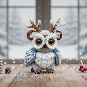 Op de afbeelding: Een handgemaakte gehaakte uil met geweien, in blauw, wit en bruin, staat op een houten oppervlak. De uil heeft grote ogen en is versierd met dennenappels en bessen. Een besneeuwd raam is op de achtergrond.