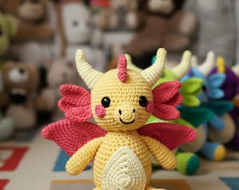May include: A handmade crochet dragon plush toy in yellow, with pink wings, horns, and accents. The dragon has a smiling face, black eyes, and rosy cheeks. Other plush toys are visible in the background.