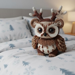 Op de afbeelding: Een gehaakte uil met grote, donkere ogen en bruine en crèmekleurige veren. De uil draagt een gewei en zit op een bed met een wit en blauw boompatroon. Een lamp en een nachtkastje staan op de achtergrond.
