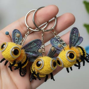 May include: Three handmade crochet bee keychains. Each bee has a yellow body with black stripes, large googly eyes, and detailed wings. The keychains are held on silver metal rings, with a blurred floral background.