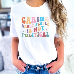 May include: White t-shirt with the text "CARING ABOUT PEOPLE IS NOT POLITICAL" in colorful, retro-style lettering. The shirt is paired with denim shorts. The overall aesthetic is casual and comfortable.