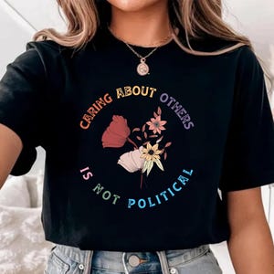 May include: Black t-shirt with the text "CARING ABOUT OTHERS IS NOT POLITICAL" in a rainbow gradient. The shirt features a floral design in shades of red, pink, yellow, and white. The word "BLACK" is printed at the bottom of the shirt.