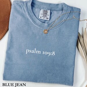 May include: A blue jean Comfort Colors t-shirt with the text "psalm 109:8" in white. A gold necklace with a pendant rests on the shirt. The shirt is folded and has a tag that reads "Comfort Colors".