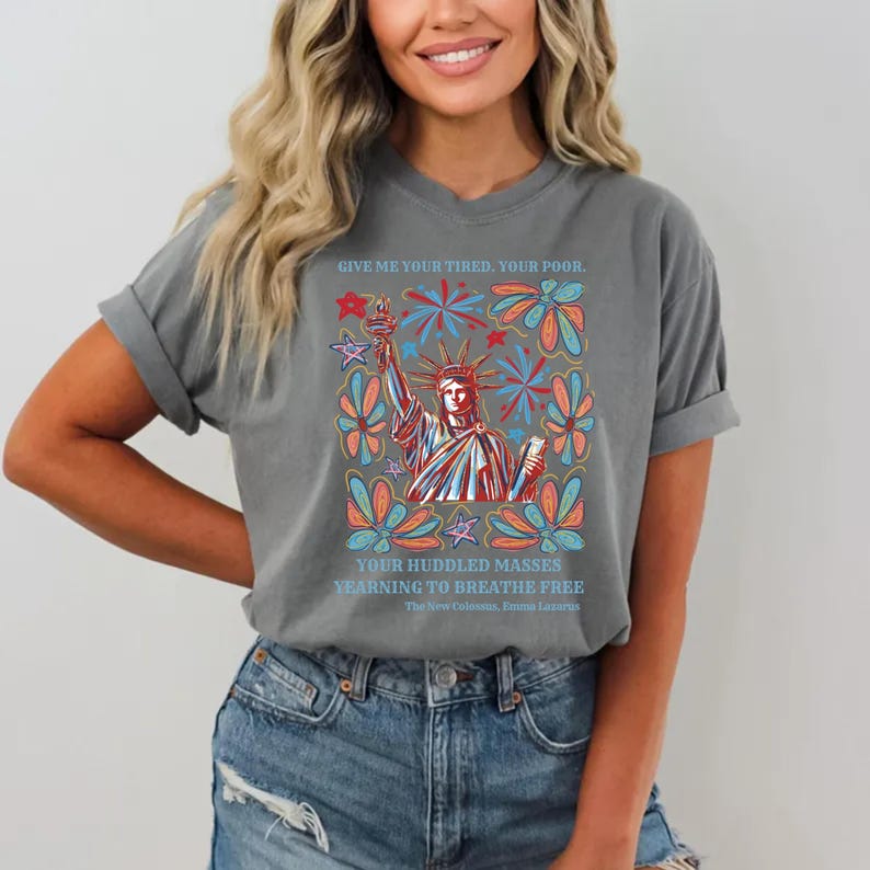 May include: Gray t-shirt featuring a colorful graphic of the Statue of Liberty with text from Emma Lazarus's poem. The design includes red, blue, and orange floral and starburst elements. The text reads "GIVE ME YOUR TIRED, YOUR POOR...YEARNING TO BREATHE FREE."