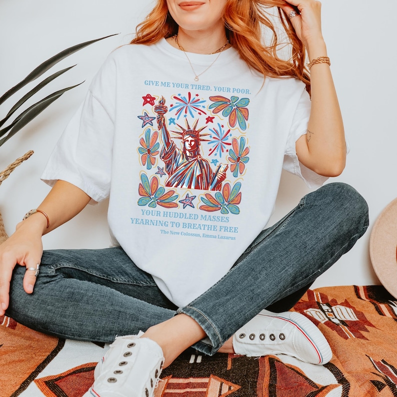 May include: White t-shirt featuring a colorful illustration of the Statue of Liberty with text that reads "GIVE ME YOUR TIRED, YOUR POOR. YOUR HUDDLED MASSES YEARNING TO BREATHE FREE." The design includes stars and floral elements.