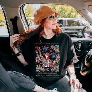 May include: Black t-shirt with a graphic of the Statue of Liberty surrounded by colorful flowers and fireworks. The text "GIVE ME YOUR TIRED. YOUR POOR. YOUR HUDDLED MASSES YEARNING TO BREATHE FREE" is printed on the shirt. The New Colossus, Emma Lazarus is also printed on the shirt.