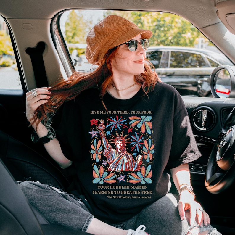 May include: Black t-shirt with a graphic of the Statue of Liberty surrounded by colorful flowers and fireworks. The text "GIVE ME YOUR TIRED. YOUR POOR. YOUR HUDDLED MASSES YEARNING TO BREATHE FREE" is printed on the shirt. The New Colossus, Emma Lazarus is also printed on the shirt.