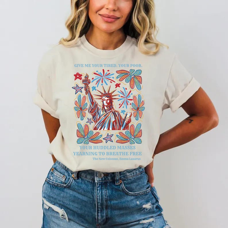 May include: Cream-colored t-shirt featuring a graphic of the Statue of Liberty with fireworks and floral designs. The text "GIVE ME YOUR TIRED, YOUR POOR" and "YOUR HUDDLED MASSES YEARNING TO BREATHE FREE" are printed above and below the image.