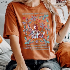 May include: Burnt orange t-shirt featuring a graphic of the Statue of Liberty holding a torch, surrounded by colorful flowers and fireworks. The text "GIVE ME YOUR TIRED, YOUR POOR" and "YOUR HUDDLED MASSES YEARNING TO BREATHE FREE" is printed above and below the image.