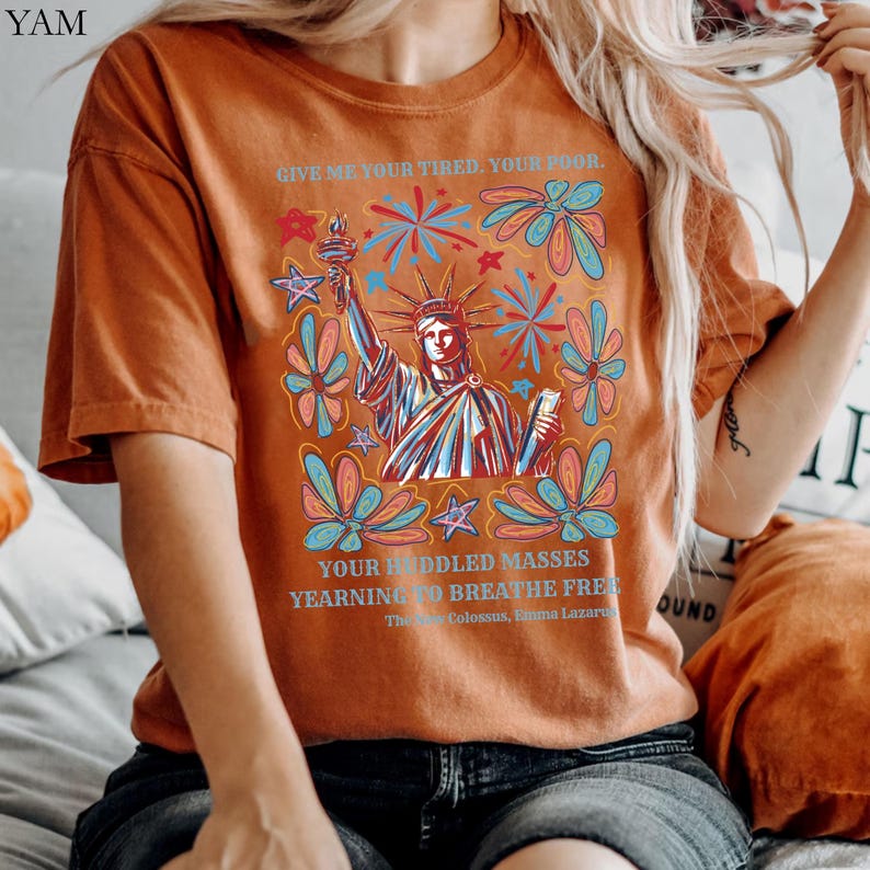 May include: Burnt orange t-shirt featuring a graphic of the Statue of Liberty holding a torch, surrounded by colorful flowers and fireworks. The text "GIVE ME YOUR TIRED, YOUR POOR" and "YOUR HUDDLED MASSES YEARNING TO BREATHE FREE" is printed above and below the image.