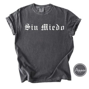 May include: Dark grey t-shirt with rolled-up sleeves. The shirt features the white text "Sin Miedo" in a gothic font. A circular logo with the word "Pepper" is visible on the bottom right.