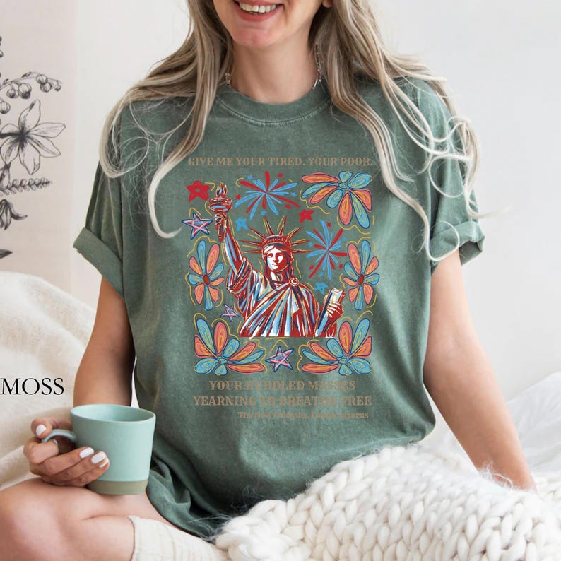 May include: Green t-shirt featuring a colorful graphic of the Statue of Liberty holding a torch, surrounded by floral and starburst designs. The text "GIVE ME YOUR TIRED, YOUR POOR..." is printed above the image, with additional text below.