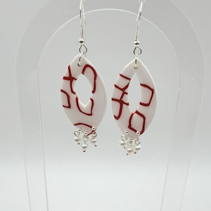 Op de afbeelding: Een paar witte, ovale oorbellen met rode abstracte patronen en kleine, bungelende parelaccenten. De oorbellen hebben zilverkleurige haken. Ze worden gepresenteerd op een transparante standaard tegen een witte achtergrond.