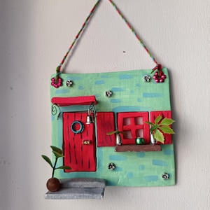 May include: A whimsical, handmade wall hanging depicting a miniature house. The house features a red door and window frames against a textured, turquoise facade. Decorative elements include a small bell, a wreath, and miniature plants.