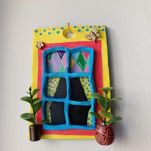 Puede incluir: Adorno de pared artesanal y colorido con diseño de ventana. La ventana tiene un marco azul, cristales negros y cortinas estampadas. Dos pequeñas plantas en macetas flanquean la ventana. Fondo amarillo con lunares azules y adornos de mariposas y flores.