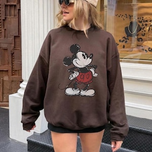 May include: A brown sweatshirt featuring a Mickey Mouse graphic. The sweatshirt has long sleeves and a crew neck. The image also shows a beige baseball cap and sunglasses.