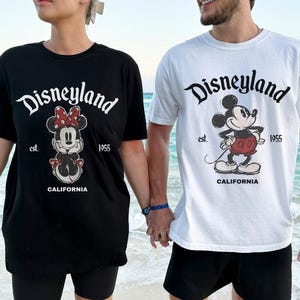 May include: Black and white Disneyland t-shirts featuring Minnie Mouse and Mickey Mouse graphics. The shirts have the Disneyland logo, the text "est. 1955", and "CALIFORNIA". The people are holding hands on a beach.