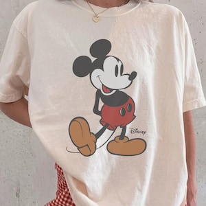 May include: An off-white short-sleeve t-shirt featuring a graphic of Mickey Mouse with the Disney logo printed in black. The character is depicted in black, white, red, and tan colors. The shirt is paired with red and white gingham patterned shorts.