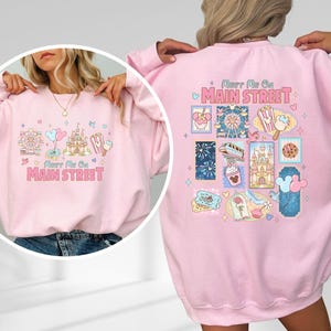 May include: A light pink sweatshirt with the text "Meet Me On Main Street" and various cartoon illustrations. The back of the sweatshirt features additional themed graphics. The sweatshirt is a casual style.