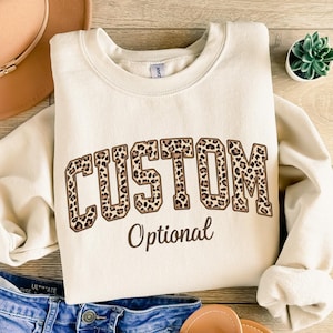 May include: A cream-coloured sweatshirt featuring the word "CUSTOM" in a leopard print design. The word "Optional" is printed below in brown script. The sweatshirt is styled with blue denim shorts, a tan hat, and sandals.