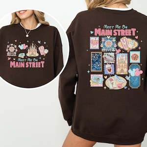 May include: A brown sweatshirt with the text "Meet Me On Main Street" in pink. The front features a Ferris wheel, castle, and popcorn graphic. The back has various themed stamp designs.