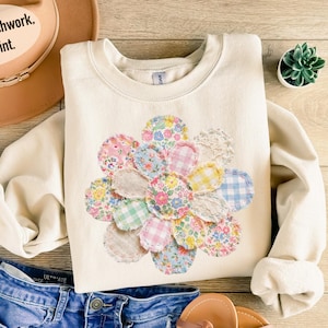 May include: A cream-coloured sweatshirt with a floral design made of colourful fabric circles. The design includes pink, blue, yellow, and green patterns. The text on the hat says "Not real patchwork. DTF print."