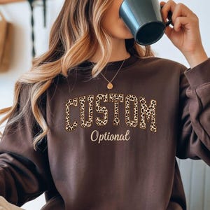 May include: A brown sweatshirt with the word "CUSTOM" in a leopard print design across the chest. Below the word "CUSTOM" is the word "Optional" in a smaller, cursive font. The sweatshirt has long sleeves and a crew neck.