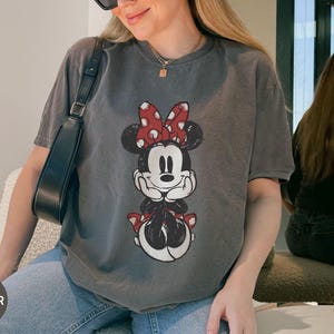 Puede incluir: Camiseta gris con un dise&ntilde;o de Minnie Mouse con un lazo rojo de lunares. La camiseta tiene un aspecto vintage y descolorido. La palabra "PEPPER" est&aacute; en un c&iacute;rculo en la esquina inferior izquierda. La camiseta se combina con vaqueros azules.