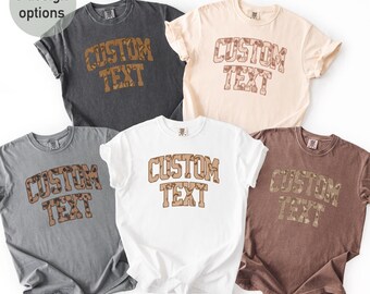 May include: Six t-shirts in various colors: charcoal, cream, light gray, white, and brown. Each shirt features the text "CUSTOM TEXT" in a brown, textured font. The shirts are short-sleeved and appear to be made of a soft material.