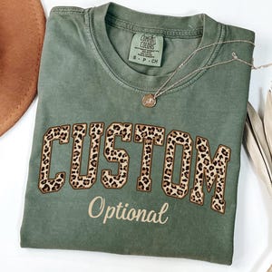 May include: An olive green t-shirt with the word "CUSTOM" in a leopard print design. The word "Optional" is below the word "CUSTOM". A silver necklace with a pendant is on the shirt.