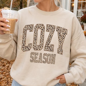 May include: A beige crewneck sweatshirt featuring the text COZY SEASON in a bold, block-letter font. The letters are filled with a brown and black leopard print pattern. The sweatshirt has long sleeves and a ribbed neckline, cuffs, and hem.