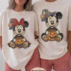 May include: Two cream-colored t-shirts featuring graphic prints of cartoon mice. One mouse wears a red bow and the other a purple witch hat, both sitting with orange jack-o'-lanterns. The designs have a textured, stitched appearance.
