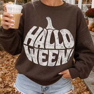 May include: A dark brown long-sleeve crewneck sweatshirt featuring a large, white, distressed-texture graphic of a pumpkin shape containing the text "HALLOWEEN!" in bold, capitalized letters across the center of the chest area.