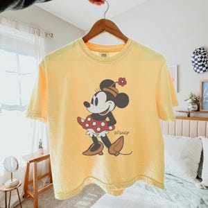May include: Yellow t-shirt featuring a Minnie Mouse graphic. The graphic shows Minnie Mouse in a red and white polka dot dress, black shoes, and a flower on her head. The Disney logo is printed below the character.