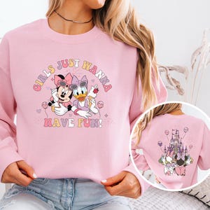 May include: A light pink sweatshirt featuring Minnie Mouse and Daisy Duck with sunglasses, holding treats, and the text "Girls Just Wanna Have Fun!". The back of the sweatshirt shows a castle and the same characters.