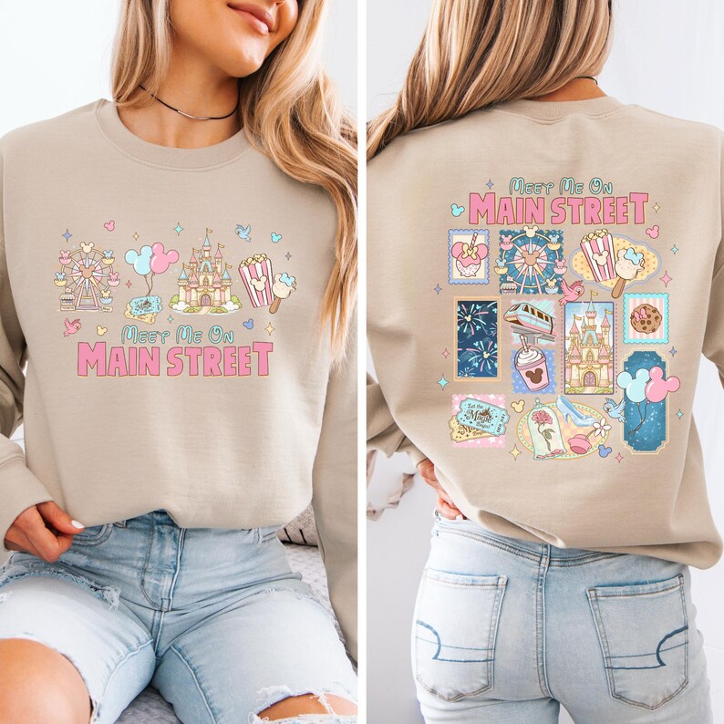 May include: Beige sweatshirt with a colorful graphic design. The front features a Ferris wheel, castle, popcorn, and ice cream, with the text "Meet Me On Main Street." The back has various themed illustrations and the same text.