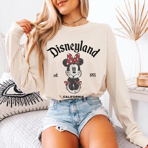 May include: Cream-colored long-sleeve shirt featuring a Minnie Mouse graphic and the text "Disneyland" above the character. The graphic includes the text "est. 1955 CALIFORNIA". The shirt is paired with denim shorts.