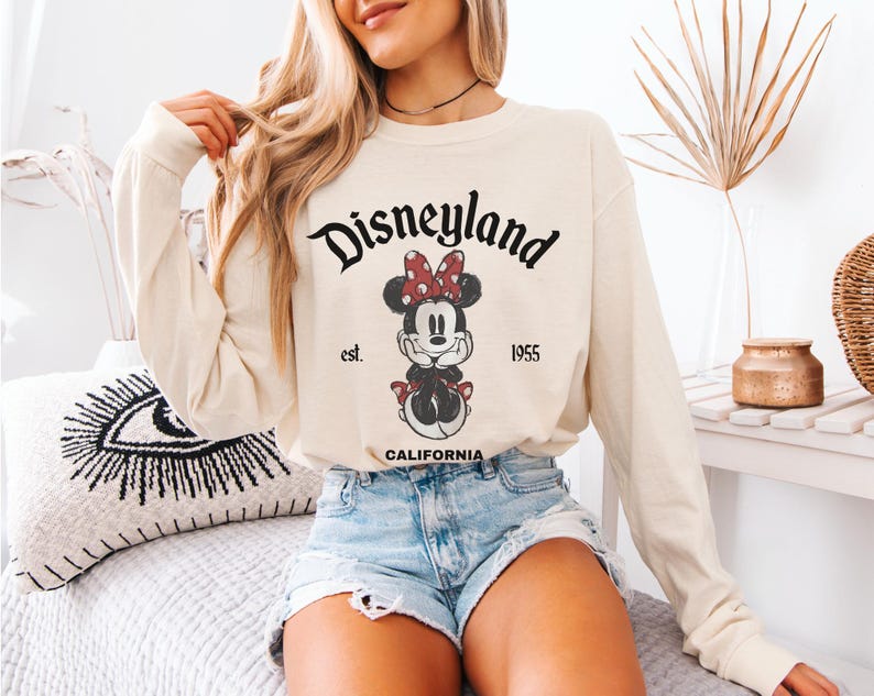 May include: Cream-colored long-sleeve shirt featuring a Minnie Mouse graphic and the text "Disneyland" above the character. The graphic includes the text "est. 1955 CALIFORNIA". The shirt is paired with denim shorts.