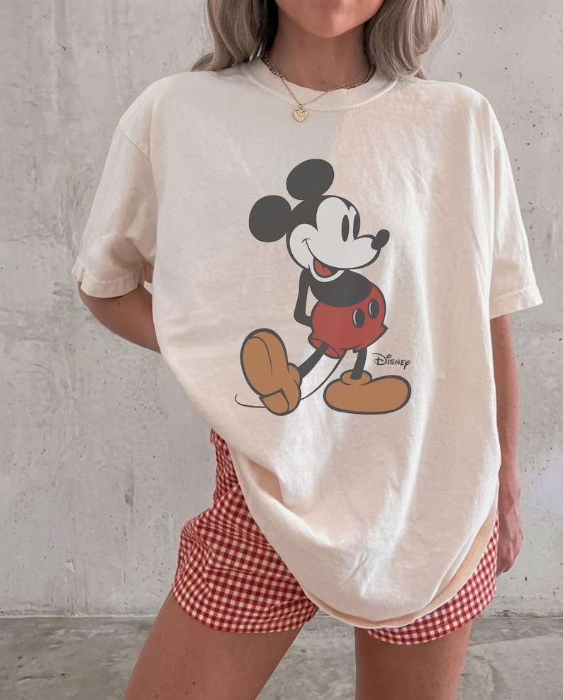 May include: An off-white short-sleeve t-shirt featuring a graphic of Mickey Mouse with the Disney logo printed in black. The character is depicted in black, white, red, and tan colors. The shirt is paired with red and white gingham patterned shorts.