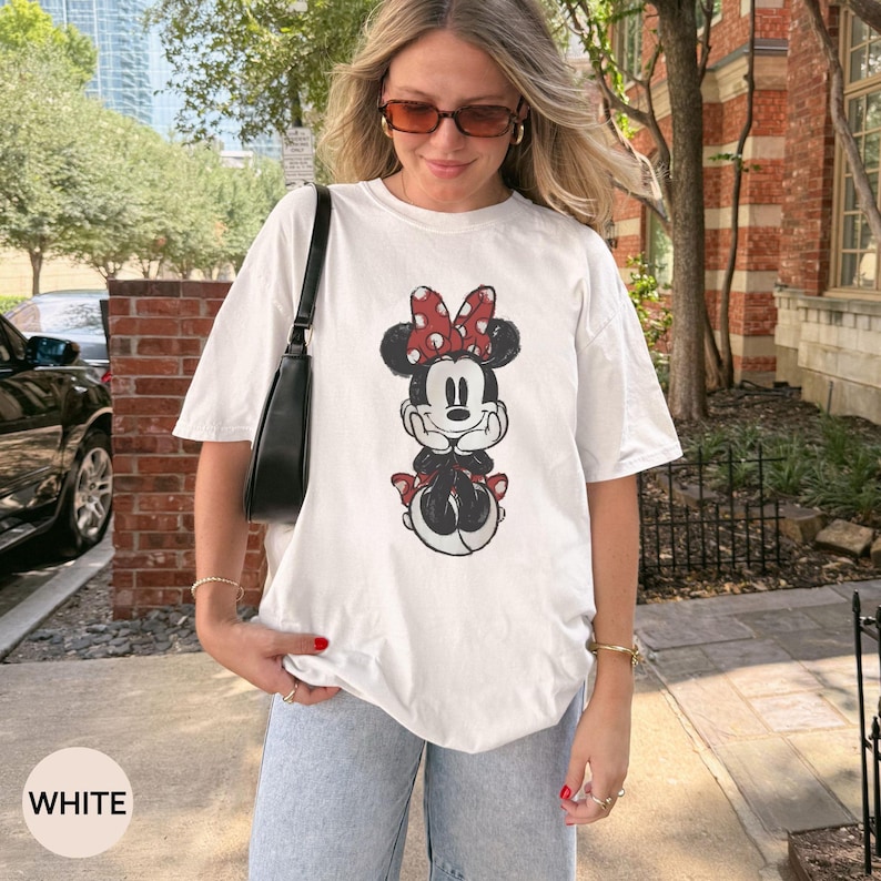 Puede incluir: Camiseta blanca con un dise&ntilde;o de Minnie Mouse con un lazo rojo de lunares. El personaje de dibujos animados est&aacute; sentado y sonriendo. La palabra "WHITE" se muestra en un rect&aacute;ngulo blanco en la esquina inferior izquierda.