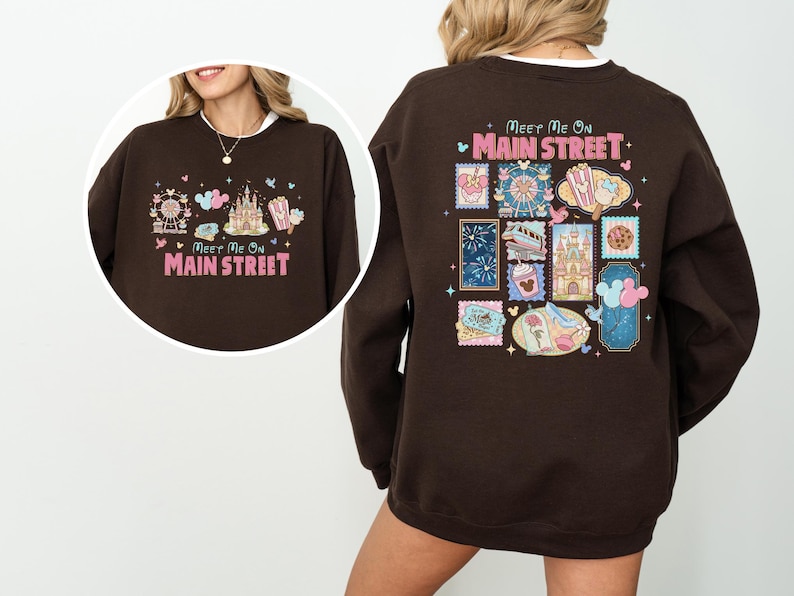 May include: A brown sweatshirt with the text "Meet Me On Main Street" in pink. The front features a Ferris wheel, castle, and popcorn graphic. The back has various themed stamp designs.