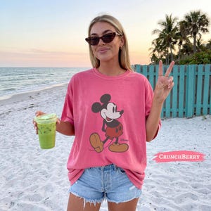 May include: A coral pink short-sleeved t-shirt featuring a graphic print of Mickey Mouse in his classic red shorts and yellow shoes. The shirt has a crew neckline and a relaxed fit, paired with light blue denim shorts with a frayed hemline.